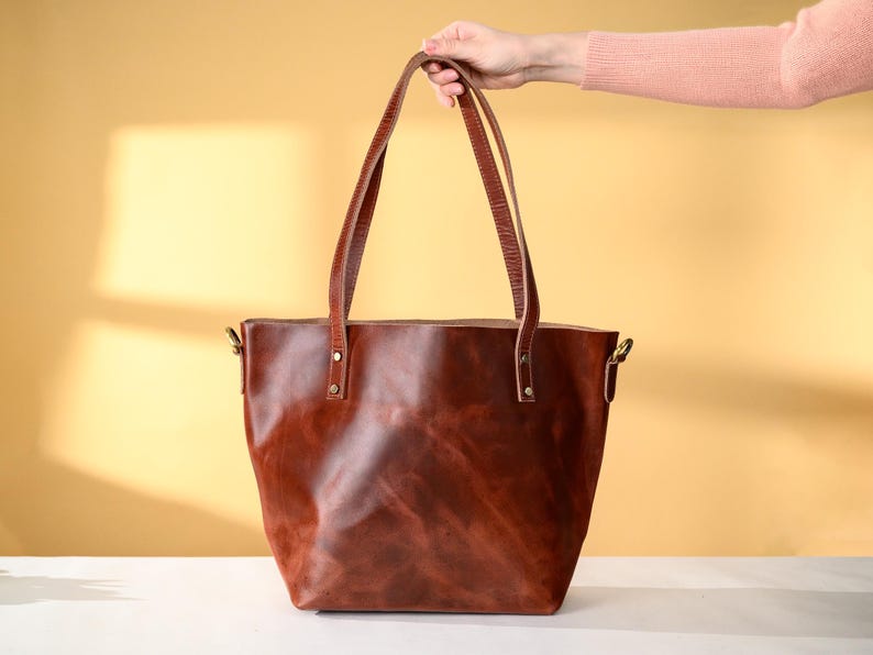Puede incluir: Un bolso tote de cuero marr&oacute;n con asas largas. El bolso es de un color marr&oacute;n rojizo intenso y parece estar hecho de cuero de alta calidad. Las asas est&aacute;n sujetas con herrajes met&aacute;licos. El bolso es sostenido por una persona.