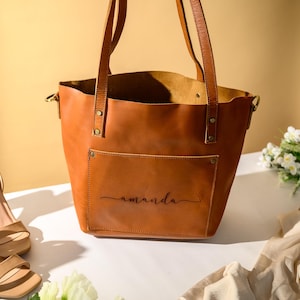 Puede incluir: Un bolso tote de cuero marr&oacute;n con un bolsillo delantero y asas. La palabra "amanda" est&aacute; escrita en el bolsillo delantero. El bolso est&aacute; junto a un par de tacones color beige y un ramo de flores blancas.