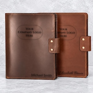 May include: Two leather portfolios in dark brown and medium brown shades. Each features a side strap with two brass-colored metal rivets, an oval engraving area with the text "YOUR COMPANY LOGO HERE," and a personalized name at the bottom.