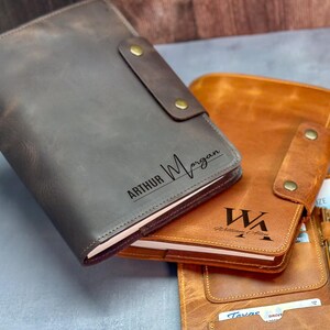 Peut inclure: Deux prot&egrave;ge-carnets en cuir avec fermetures &agrave; pression. La couverture gris fonc&eacute; est grav&eacute;e du nom Arthur Morgan. La couverture beige est grav&eacute;e des initiales WA et du nom William Allen. Un stylo dor&eacute; et des cartes sont ins&eacute;r&eacute;s dans la couverture.