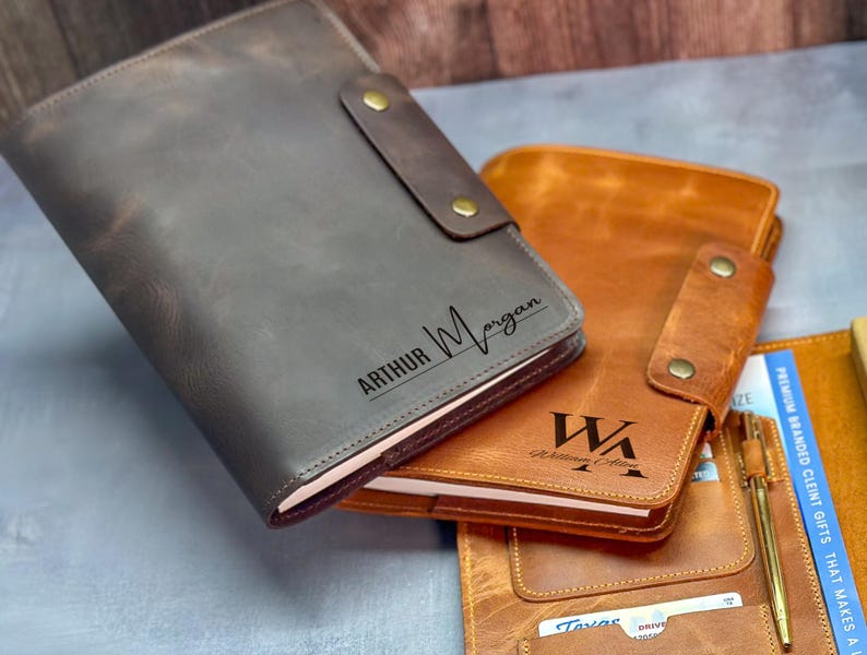 Peut inclure: Deux prot&egrave;ge-carnets en cuir avec fermetures &agrave; pression. La couverture gris fonc&eacute; est grav&eacute;e du nom Arthur Morgan. La couverture beige est grav&eacute;e des initiales WA et du nom William Allen. Un stylo dor&eacute; et des cartes sont ins&eacute;r&eacute;s dans la couverture.