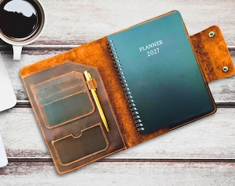 May include: A brown leather planner cover featuring a dark green spiral-bound 2027 planner inside. The cover includes multiple interior pockets for cards and a gold pen. A snap closure secures the flap on the right side of the organizer.