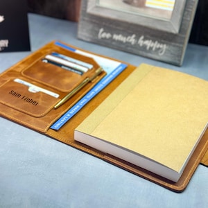 May include: A brown leather notebook cover with a pen, cards, and the name "Sam Fisher" embossed on the front. The cover is open to reveal a blank notebook.