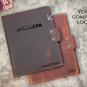 May include: Two leather document holders, one dark brown and one reddish-brown, are displayed. The dark brown holder has "Krause CPA" and "Robert Brown" printed on it. The reddish-brown holder has "KSN" printed on it. Both have a leather strap closure.