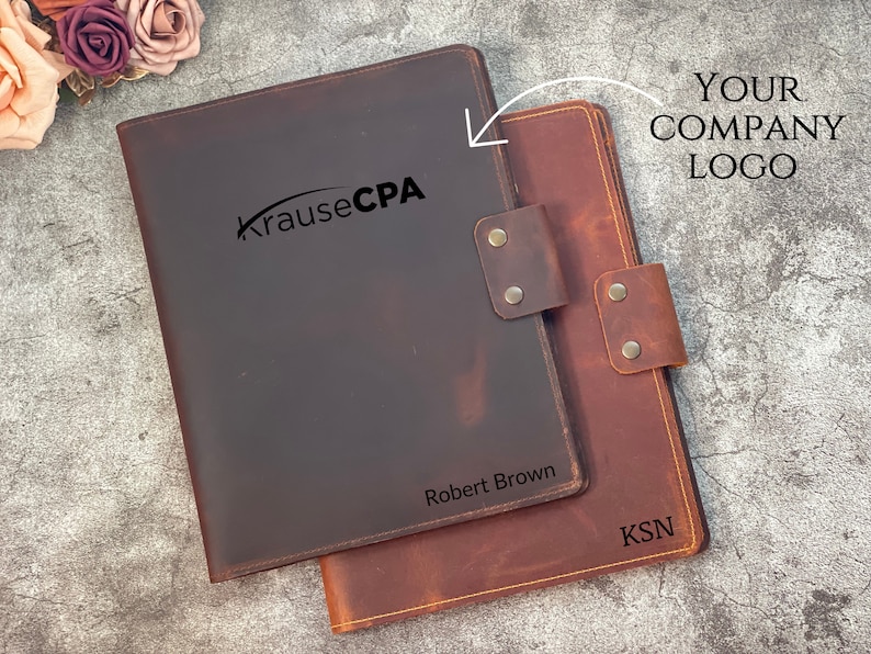 May include: Two leather document holders, one dark brown and one reddish-brown, are displayed. The dark brown holder has "Krause CPA" and "Robert Brown" printed on it. The reddish-brown holder has "KSN" printed on it. Both have a leather strap closure.