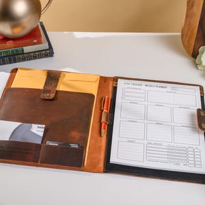 May include: A brown leather portfolio with a weekly planner, pen, and card slots. The planner is open, displaying a schedule layout with the text "Stay Focused Weekly Planner." The portfolio has a leather strap closure and a yellow envelope inside.