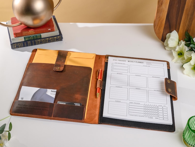 May include: A brown leather portfolio with a weekly planner, pen, and card slots. The planner is open, displaying a schedule layout with the text "Stay Focused Weekly Planner." The portfolio has a leather strap closure and a yellow envelope inside.