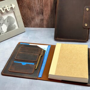Peut inclure: Une couverture de carnet en cuir marron fonc&eacute; pr&eacute;sentant un int&eacute;rieur avec plusieurs fentes pour cartes, un porte-stylo avec un stylo dor&eacute; et un carnet en papier beige clair. Une seconde couverture ferm&eacute;e avec deux boutons-pression en laiton.