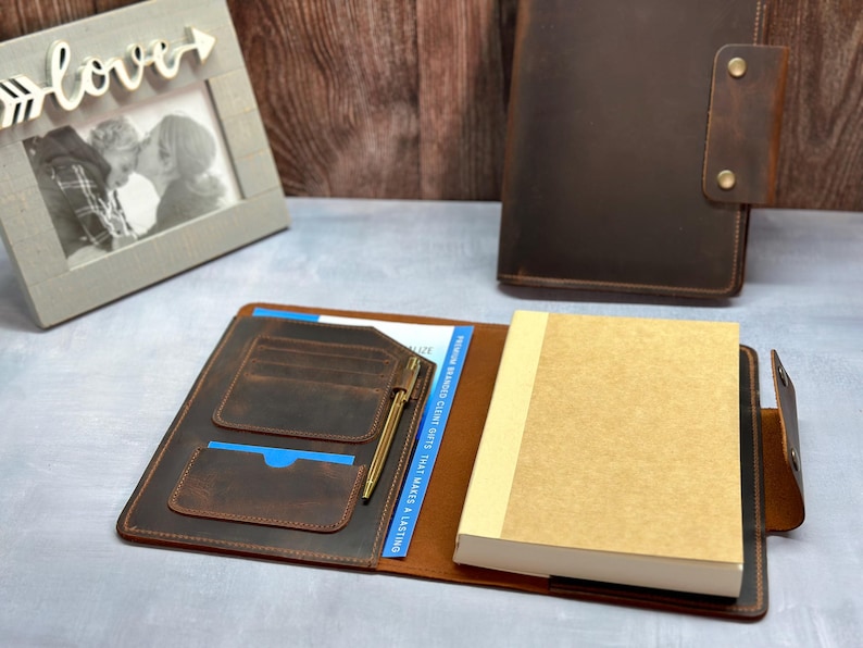Peut inclure: Une couverture de carnet en cuir marron fonc&eacute; pr&eacute;sentant un int&eacute;rieur avec plusieurs fentes pour cartes, un porte-stylo avec un stylo dor&eacute; et un carnet en papier beige clair. Une seconde couverture ferm&eacute;e avec deux boutons-pression en laiton.