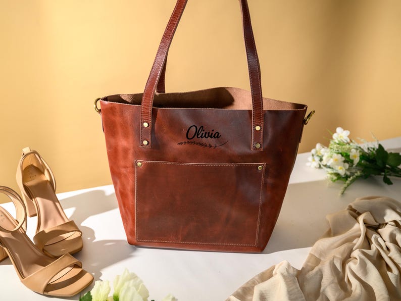 Puede incluir: Un bolso tote de cuero marr&oacute;n con un bolsillo delantero y asas largas. La bolsa tiene el nombre "Olivia" impreso. La bolsa est&aacute; al lado de un par de sandalias beige de tac&oacute;n alto y un ramo de flores blancas.