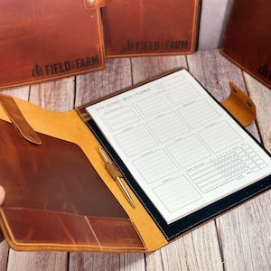 May include: A brown leather planner with a weekly layout, pen, and pocket. The planner is open, displaying the "Stay Focused - Weekly Planner" text. The cover has the "FIELD & FARM" logo. Additional planners are visible in the background.
