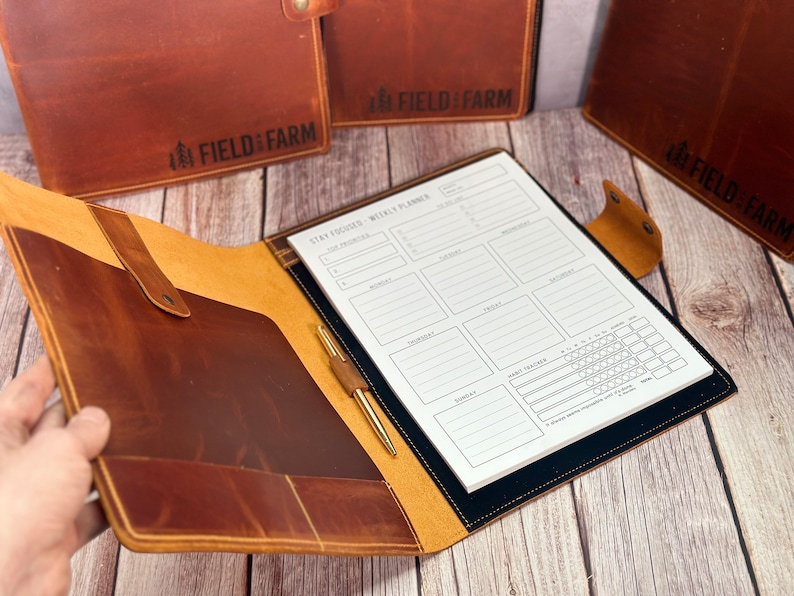 May include: A brown leather planner with a weekly layout, pen, and pocket. The planner is open, displaying the "Stay Focused - Weekly Planner" text. The cover has the "FIELD & FARM" logo. Additional planners are visible in the background.