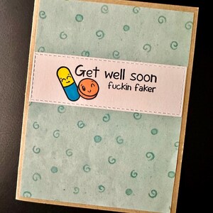 May include: A light blue card with a white border and a light brown border. The card has a green background with white circles and swirls. The card has a white banner with the text "Get well soon fuckin faker" and a cartoon image of two pills with faces.