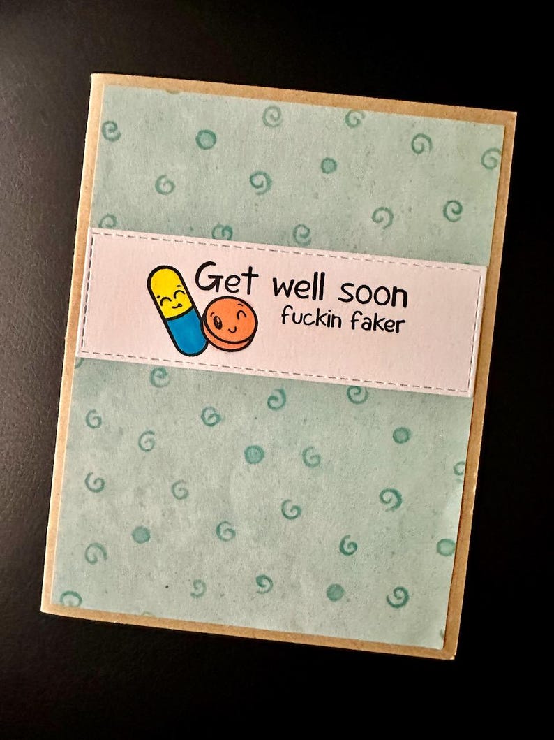 May include: A light blue card with a white border and a light brown border. The card has a green background with white circles and swirls. The card has a white banner with the text "Get well soon fuckin faker" and a cartoon image of two pills with faces.