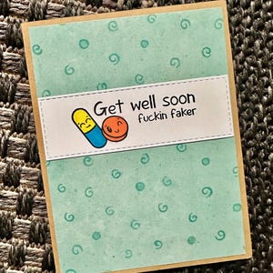 May include: A light blue card with a white border and a white rectangle in the center. The rectangle has a cartoon image of two pills with faces and the text "Get well soon fuckin faker". The card has a light blue background with a pattern of small circles and swirls.