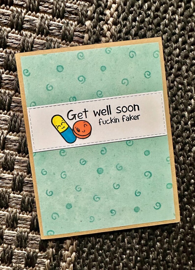 May include: A light blue card with a white border and a white rectangle in the center. The rectangle has a cartoon image of two pills with faces and the text "Get well soon fuckin faker". The card has a light blue background with a pattern of small circles and swirls.