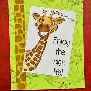 May include: A humorous greeting card with a cartoon giraffe with large eyes and a wide smile. The card has a green background with a pattern of marijuana leaves and a yellow giraffe pattern on the side. The text on the card reads "It's Your Day Enjoy the high life!"