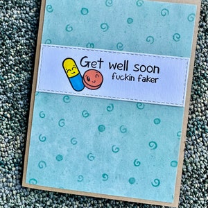 May include: A light blue card with a white panel featuring a cartoon pill and a red circle with a smiley face. The text on the card reads "Get well soon fuckin faker".