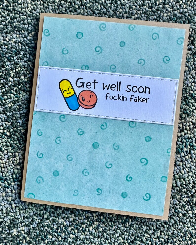 May include: A light blue card with a white panel featuring a cartoon pill and a red circle with a smiley face. The text on the card reads "Get well soon fuckin faker".