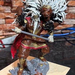 Puede incluir: Una figurita miniatura pintada a mano de un guerrero con un tocado de plumas blancas, una capa roja y dorada y una espada. La figurita está de pie sobre una base gris y marrón.