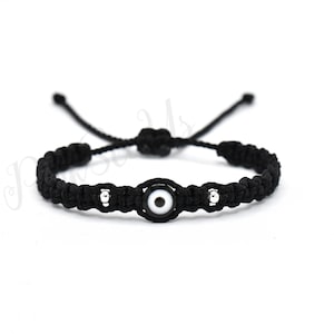 May include: Black macrame bracelet featuring an evil eye charm. The bracelet has a woven design with a central white and black eye-shaped bead, flanked by silver-colored beads. Adjustable sliding knot closure.