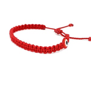 May include: A red braided cord bracelet featuring a central circular evil eye charm with a white, black, and blue iris. The bracelet includes an adjustable sliding knot closure with two thin red cord ends finished with small red beads at the tips.