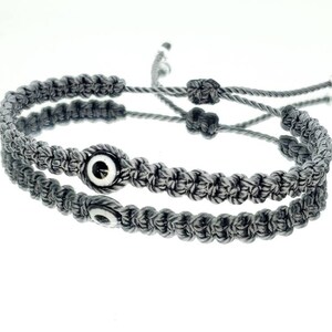 May include: Two gray braided bracelets with silver evil eye charms. The bracelets are tied together with a knot.