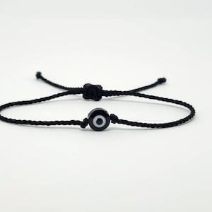 May include: A black string bracelet with a small black evil eye charm. The charm has a white center and a black pupil.