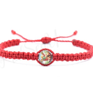 May include: Red macrame bracelet with an adjustable sliding knot closure. The bracelet features a central round charm with a colorful image. The bracelet is made of woven red cord.