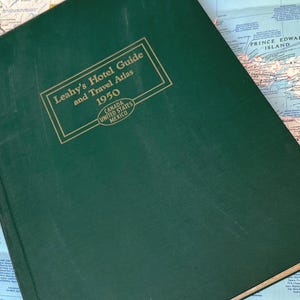 以下が含まれることがあります： 1950年のヴィンテージグリーンのLeahy's Hotel Guide and Travel Atlas。本の表紙には金色の文字と長方形のデザインが施されています。アトラスは、カナダ、米国、メキシコを含む北米の地図上に開かれています。