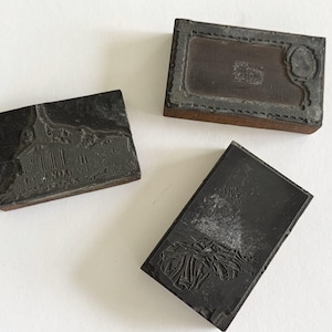 May include: Three vintage printing blocks with detailed designs. One block features a building, another a decorative frame, and the third a floral pattern. The blocks are dark gray and rectangular, with visible wear and tear, suggesting age and use.