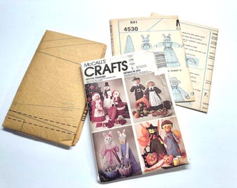 Può includere: Tre buste di cartamodelli McCall's con illustrazioni di bambole artigianali e decorazioni festive. La busta anteriore mostra il testo McCall's Crafts Festive Touches, che presenta varie figure in tessuto tra cui conigli, pupazzi di neve e personaggi stagionali in abiti colorati.