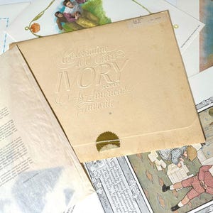 Puede incluir: Un anuncio vintage de jabón Ivory. Una carpeta beige con texto en relieve que dice "Celebrating 100 Years Ivory Soap As America's Favorite". La carpeta está abierta, revelando materiales impresos e ilustraciones.