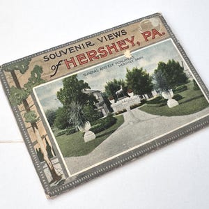 May include: Vintage souvenir booklet titled "Souvenir Views of Hershey, PA." The cover features a color image of Hershey Park's Sundial and Elk Monument. The booklet has a decorative border and is slightly worn.