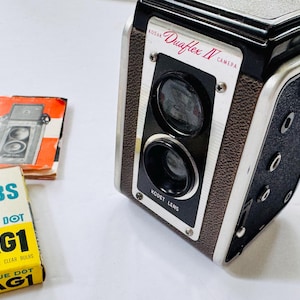 May include: A vintage Kodak Duaflex IV camera with a black and brown textured body, featuring a Kodet lens and dual lens system. Beside it lies an orange instruction manual and a yellow box labeled AG1 Blue Dot twelve clear bulbs.