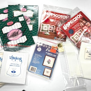 May include: A collection of cross-stitch fabric packages from brands including Charles Craft, Hopscotch, White Horse's, and Zweigart. The items are packaged in clear plastic bags, featuring various labels, text, and printed images of finished cross-stitch projects.