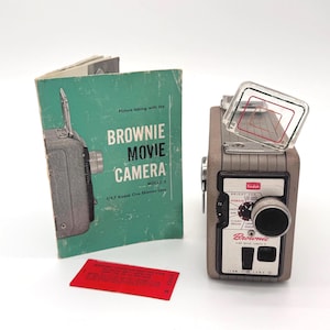 May include: A gray Kodak Brownie movie camera model 2 with a clear viewfinder, a teal instruction manual titled Picture taking with the BROWNIE MOVIE CAMERA, and a small red rectangular card with printed text.