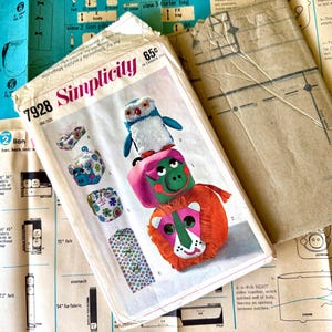 May include: A Simplicity 7928 sewing pattern envelope featuring illustrations of a stacked owl, pig, and lion toy, along with a tissue box cover and garment bag. The envelope is printed with the text Simplicity 7928 and 65c in Canada.