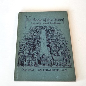 Puede incluir: Un libro antiguo titulado "The Book of the Street" de Lowrie y Ludlum. La portada es de color verde azulado apagado con una ilustración de una escena callejera. También se ve el texto "High Street" Old Philadelphia 1776.