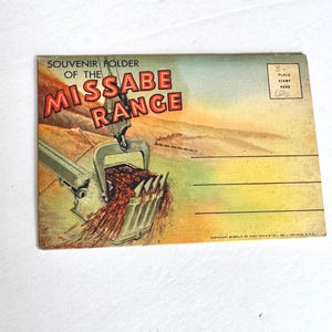 以下が含まれることがあります： 「Souvenir Folder of the Missabe Range」と題されたヴィンテージポストカード。鉱山用掘削機が資材を投棄するイラストが特徴です。テキストは赤色で、背景は黄色とオレンジ色の空のある風景です。
