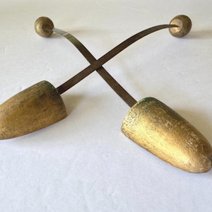 Puede incluir: Dos hormas de zapatos antiguos. Cada horma tiene una pieza de puntera redondeada de color dorado, un soporte metálico curvado y un pequeño pomo redondo. Las hormas están cruzadas sobre un fondo blanco.