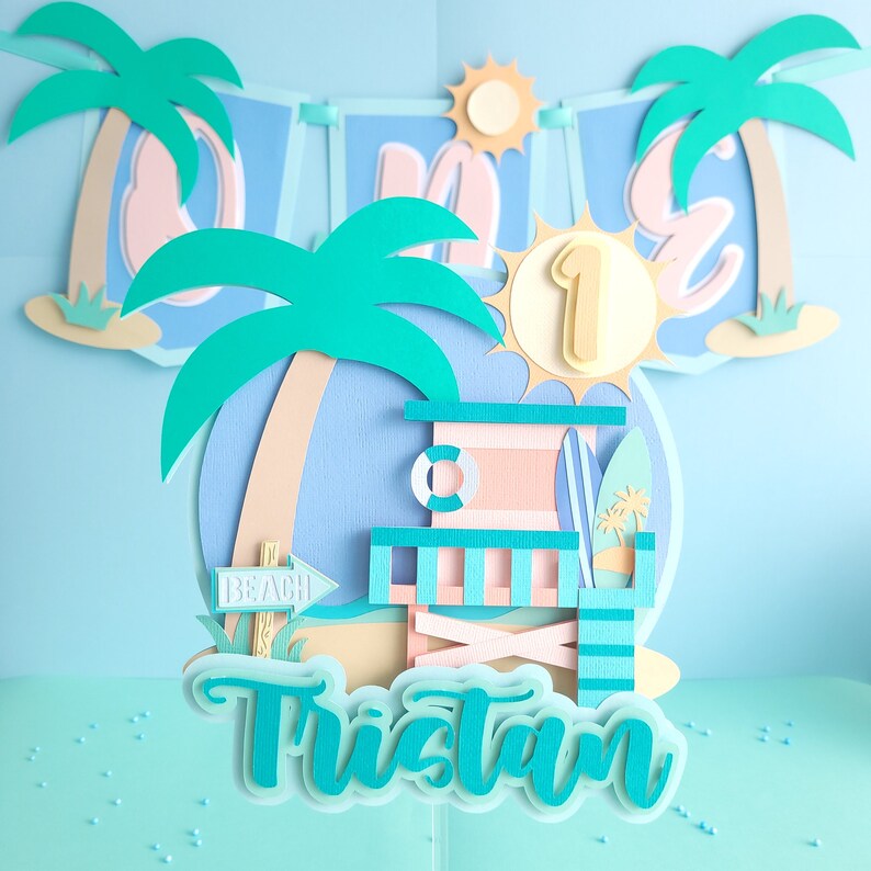 May include: A turquoise blue and white paper cake topper with a beach theme. The topper features a palm tree, a lifeguard stand, a surfboard, and the words "Beach" and "Tristan".