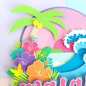 May include: Paper craft featuring a tropical scene with a palm tree, hibiscus flowers, and a blue wave. The scene is set against a pink and purple background.