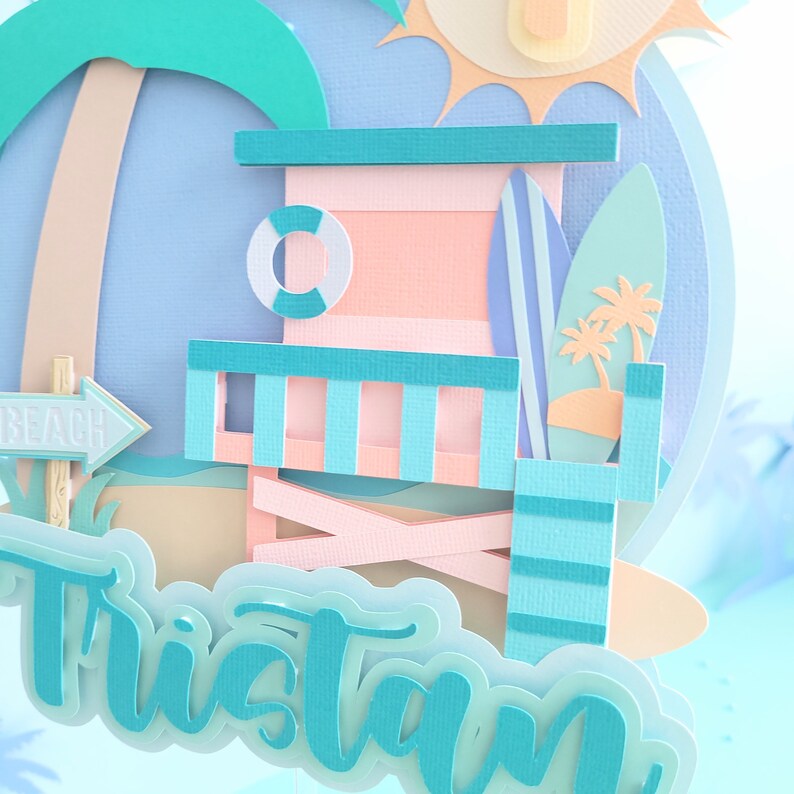 May include: A turquoise and pink papercraft beach scene with a lifeguard stand, palm trees, surfboards, and the name Tristan.