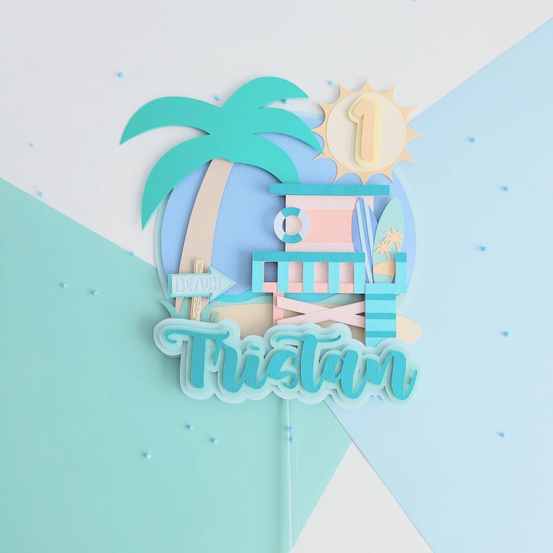 May include: A blue and pink beach-themed cake topper with a palm tree, lifeguard stand, surfboard, and the name Tristan. The sun is yellow with the number 1 in the center.