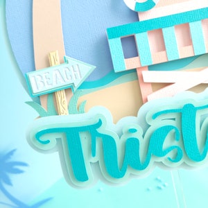 May include: A turquoise blue papercraft cake topper with the name "Trist" in a whimsical font. The topper features a beach scene with a lifeguard stand, a palm tree, and a sign that says "BEACH".