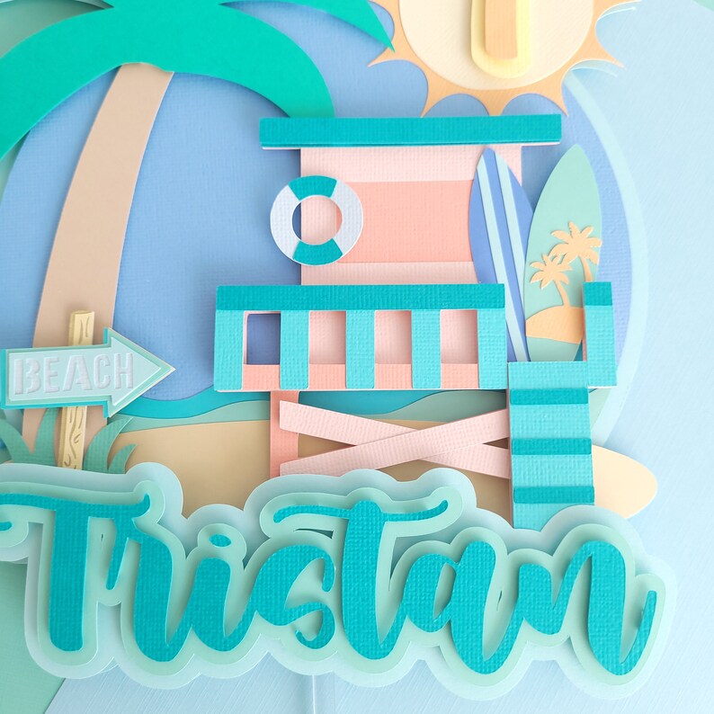 May include: A blue and green papercraft name banner with the name "Tristan" in a whimsical font. The banner features a beach scene with a lifeguard stand, palm tree, surfboard, and a sign that says "Beach".