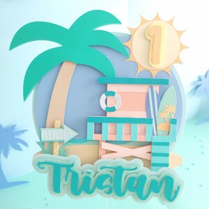 May include: A turquoise blue and pink beach hut cake topper with a palm tree, surfboard, and a sign that says "Beach". The sun is yellow with a number 1 in the center. The name Tristan is written in turquoise blue letters.