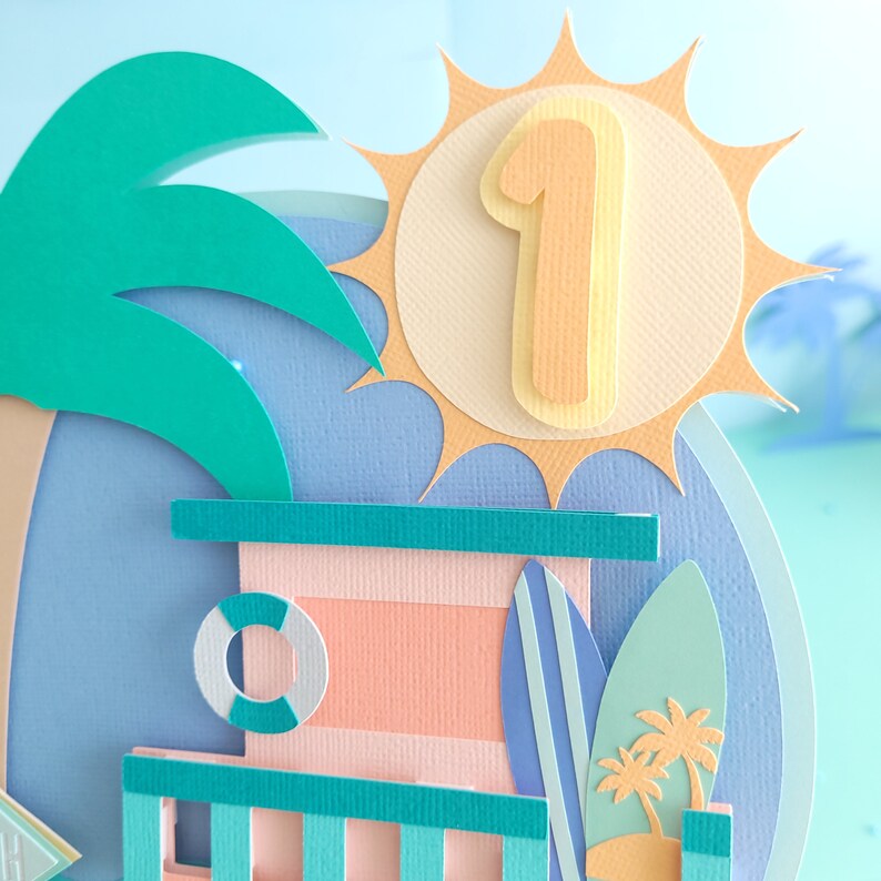 May include: A papercraft scene with a turquoise blue palm tree, a light blue and pink beach hut, a yellow sun with the number "1" on it, and two turquoise blue surfboards.