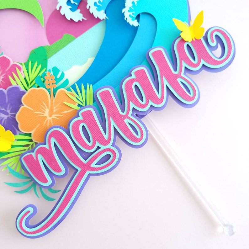 May include: A pink and blue cake topper with the name "Malaya" written in a cursive font. The topper is decorated with tropical flowers and a blue wave.